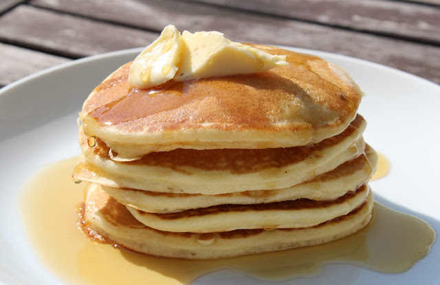 Mein originales Bild von Pancakes aus dem Mai 2011. Ein Stapel Pancakes mit Butter und Ahornsirup in einer Nahaufnahme auf einem weißen Teller in der Sonne auf einem braunen Gartentisch | moeyskitchen.com