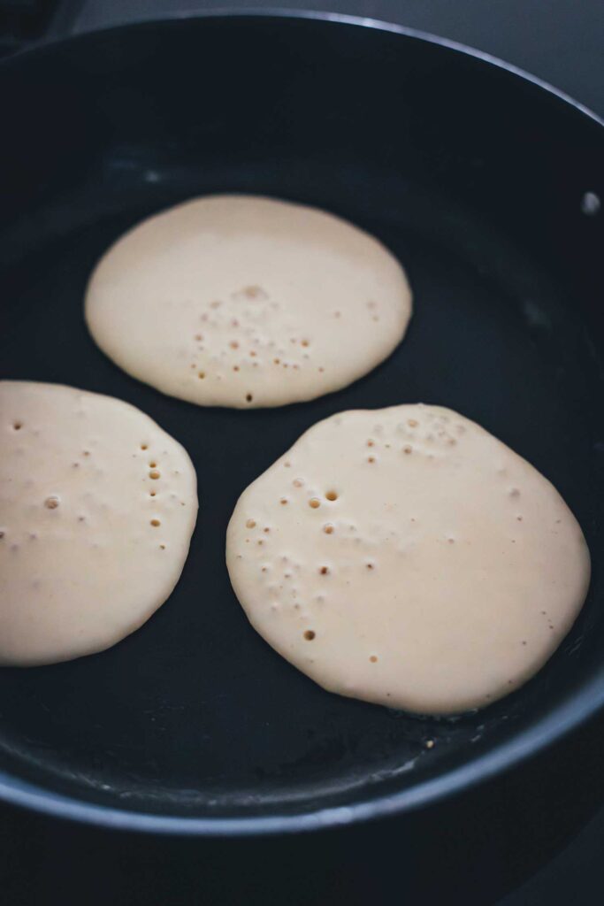 3 Pancakes bei der Zubereitung in der Pfanne – die Oberfläche wirft Blasen, bereit zum Wenden | moeyskitchen.com