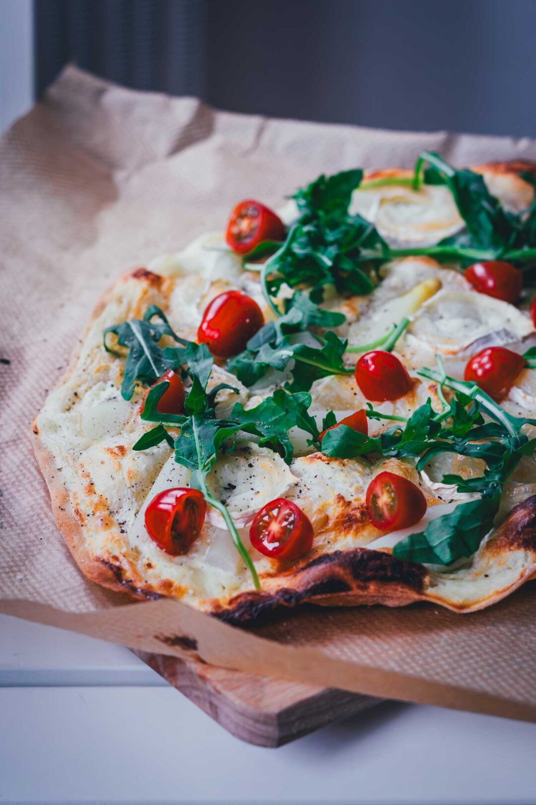 Seitliche Ansicht auf einen frisch gebackenen, knusprigen Flammkuchen mit Spargel. Zusätzlich belegt mit kleinen Tomaten, Rucola und Ziegenkäserolle perfekt für den Frühling!