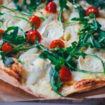 Nahaufnahme von einem knusprigen Spargel-Flammkuchen, der mit weißem Spargel, Rucola, Tomaten und Ziegenkäse belegt ist.