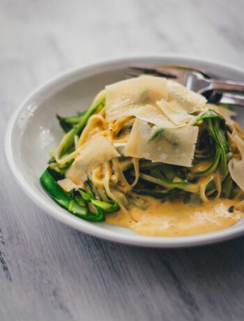 Pasta mit grünem Spargel und Parmesansauce auf einem Teller mit Parmesanspänen serviert, seitlich zu sehen.