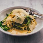 Pasta mit grünem Spargel und creiger Parmesansauce auf einem weißen Teller, garniert mit Parmesanspänen und frisch gemahlenem Pfeffer