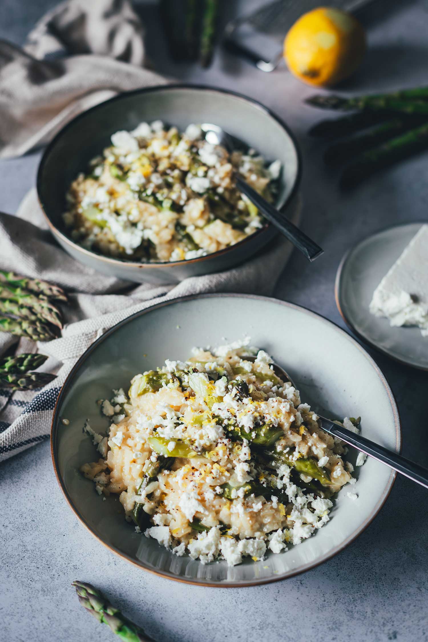 Zwei Schalen Ofenrisotto mit grünem Spargel und Feta, daneben frische Spargelstangen und ein Stück Schafsmilch-Feta