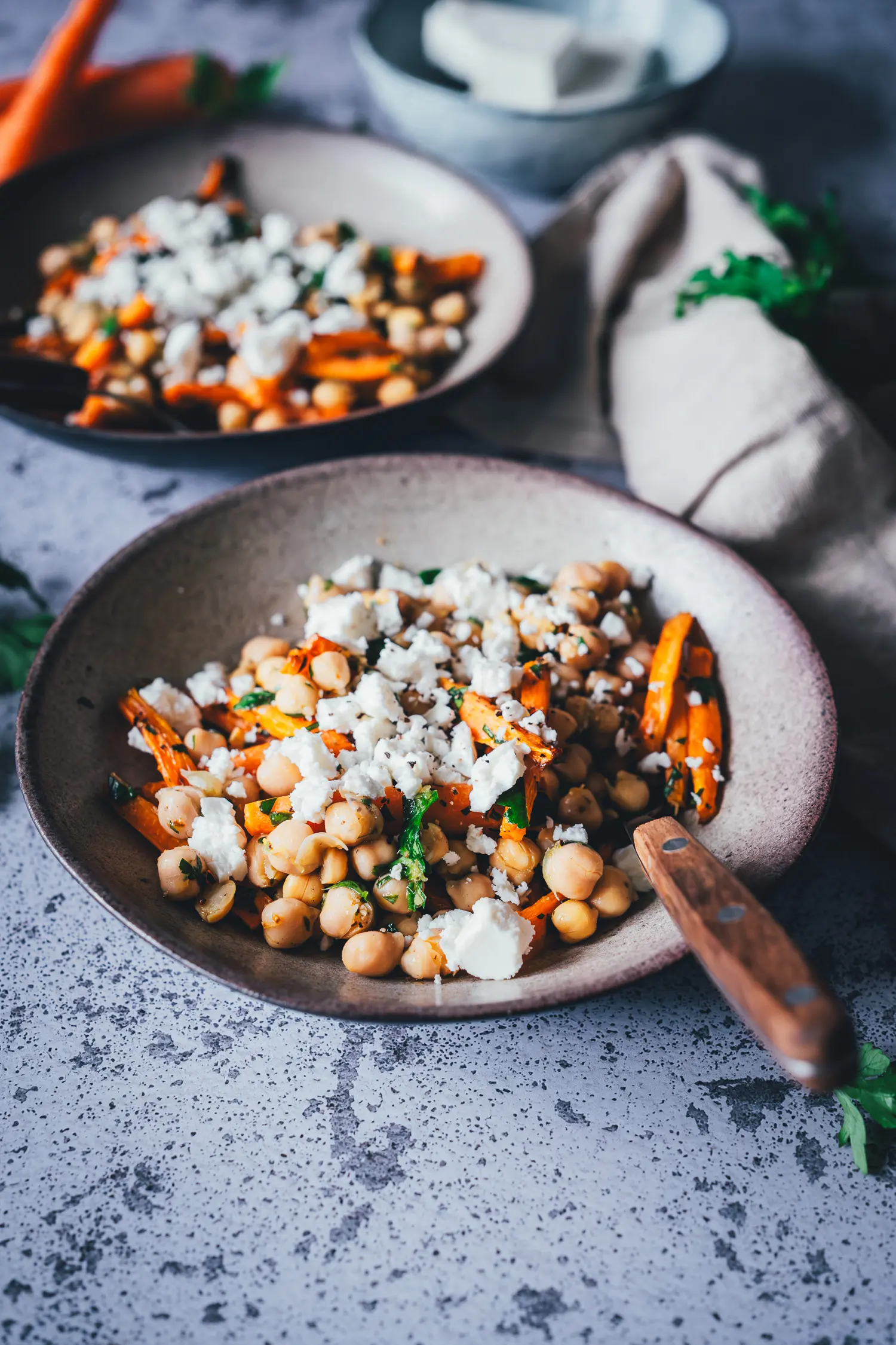 Zwei Schalen Kichererbsen-Salat mit gerösteten Möhren und Feta auf grau gesprenkeltem Untergrund, dahinter eine kleine Schale Feta