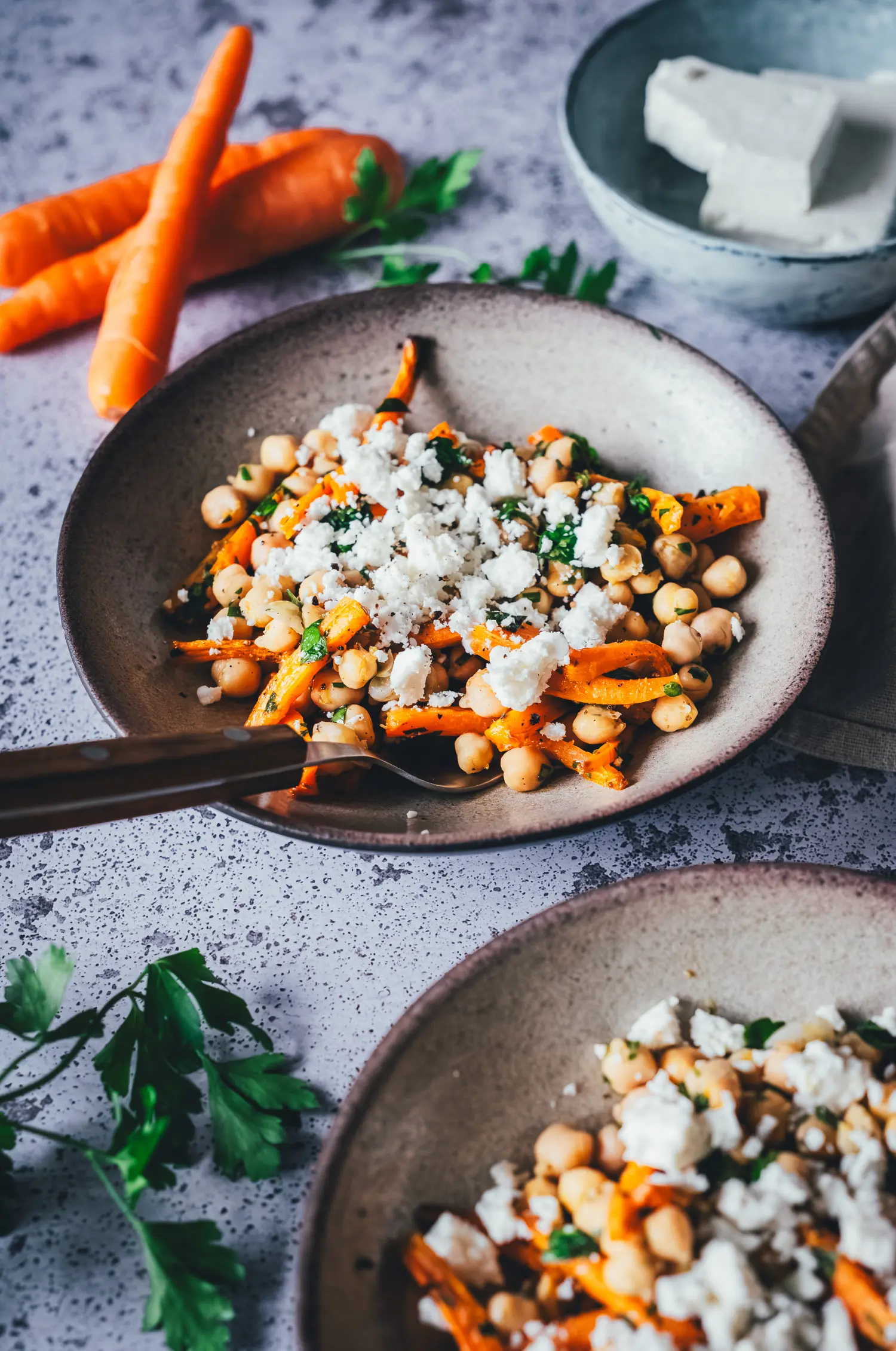 Kichererbsen-Salat mit gerösteten Möhrenstiften und Feta in einer Schale, im Hintergrund rohe Bundmöhren und ein Block Feta