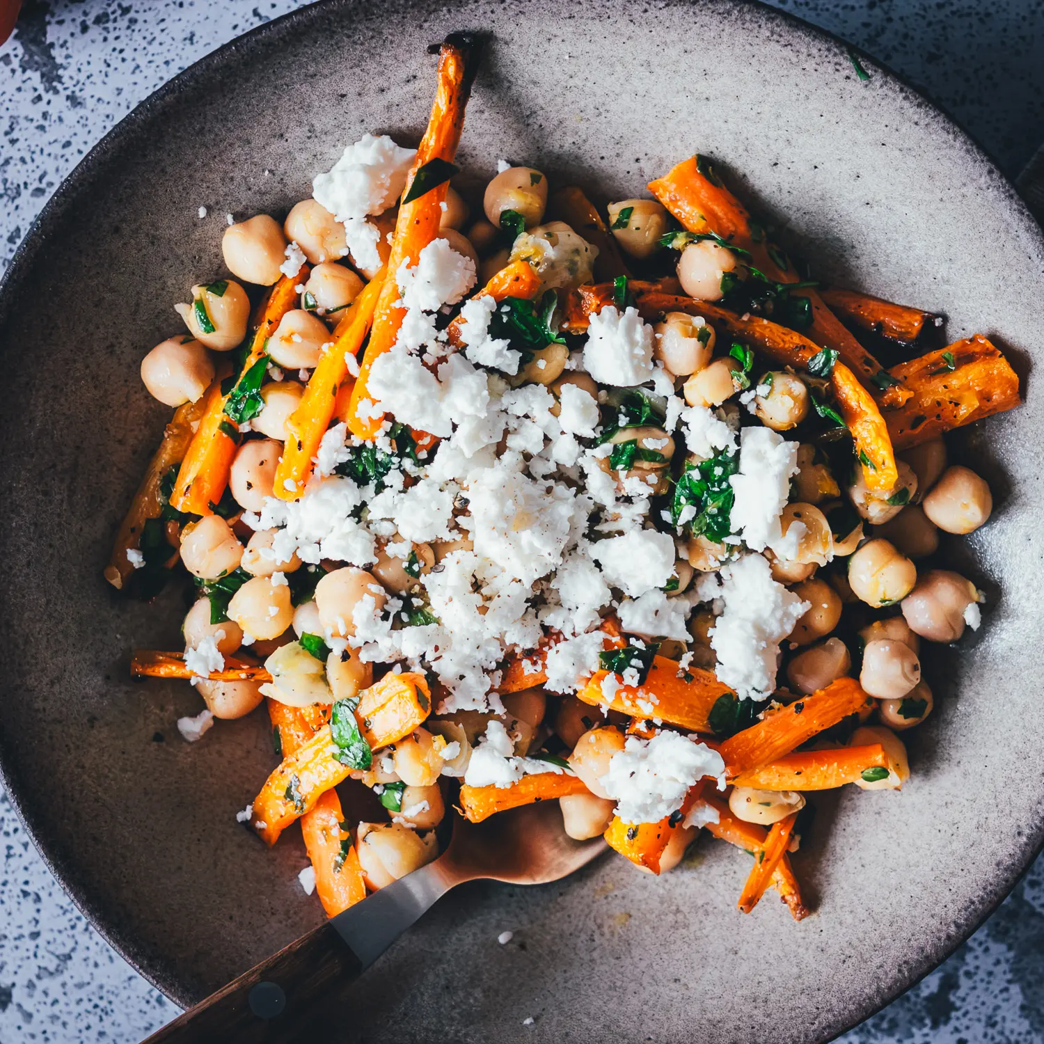 Schale Kichererbsen-Salat mit gerösteten Möhren, Feta und frischer Petersilie mit Holzgabel, umgeben von rohen Möhren und Feta