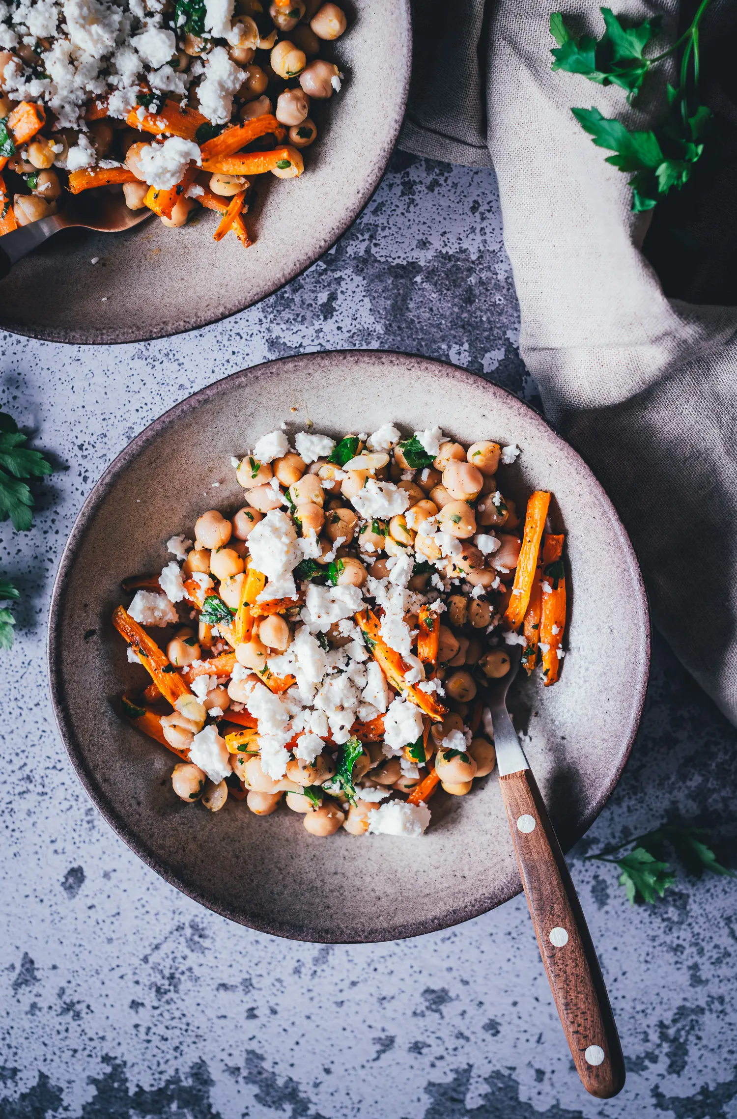 Zwei Schüsseln Möhren-Kichererbsen-Salat mit zerbröckeltem Feta von oben fotografiert, mit Leinenserviette und Petersilie