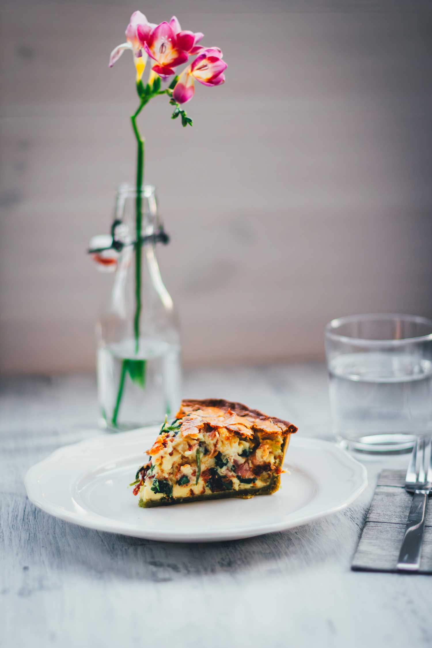 Frontale Draufsicht auf ein einzelnes Stücke Quiche auf einem weißen Teller. Daneben steht eine einzelne Blume in einer kleinen Flasche. Im Vordergrund sieht man Besteck und eine Serviette, im Hintergrund ein Glas Wasser.