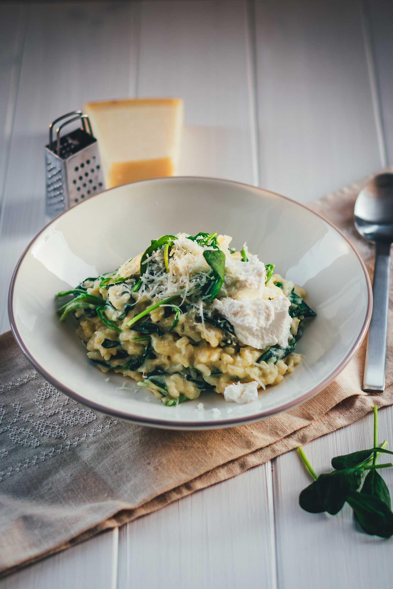 Man sieht einen hellgrauen Teller, der mit cremigem Ofenrisotto mit Spinat gefüllt ist. Daneben liegen Löffel, Parmesan und Spinat. Das Gericht sieht sehr cremig aus und wir mit Käse getoppt, bereit zum Servieren.