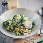 Man sieht einen hellgrauen Teller, der mit cremigem Ofenrisotto mit Spinat gefüllt ist. Daneben liegen Löffel, Parmesan und Spinat. Das Gericht sieht sehr cremig aus und wir mit Käse getoppt, bereit zum Servieren.