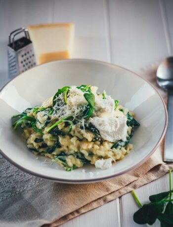Man sieht einen hellgrauen Teller, der mit cremigem Ofenrisotto mit Spinat gefüllt ist. Daneben liegen Löffel, Parmesan und Spinat. Das Gericht sieht sehr cremig aus und wir mit Käse getoppt, bereit zum Servieren.