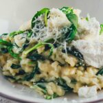 Detaillierte Nahaufnahme eines tiefen Tellers, gefüllt mit cremigem Ofenrisotto mit Spinat. Mit Ricotta und Parmesan bestreut serviert, sieht man, wie cremig und aroamtisch das Gericht ist.
