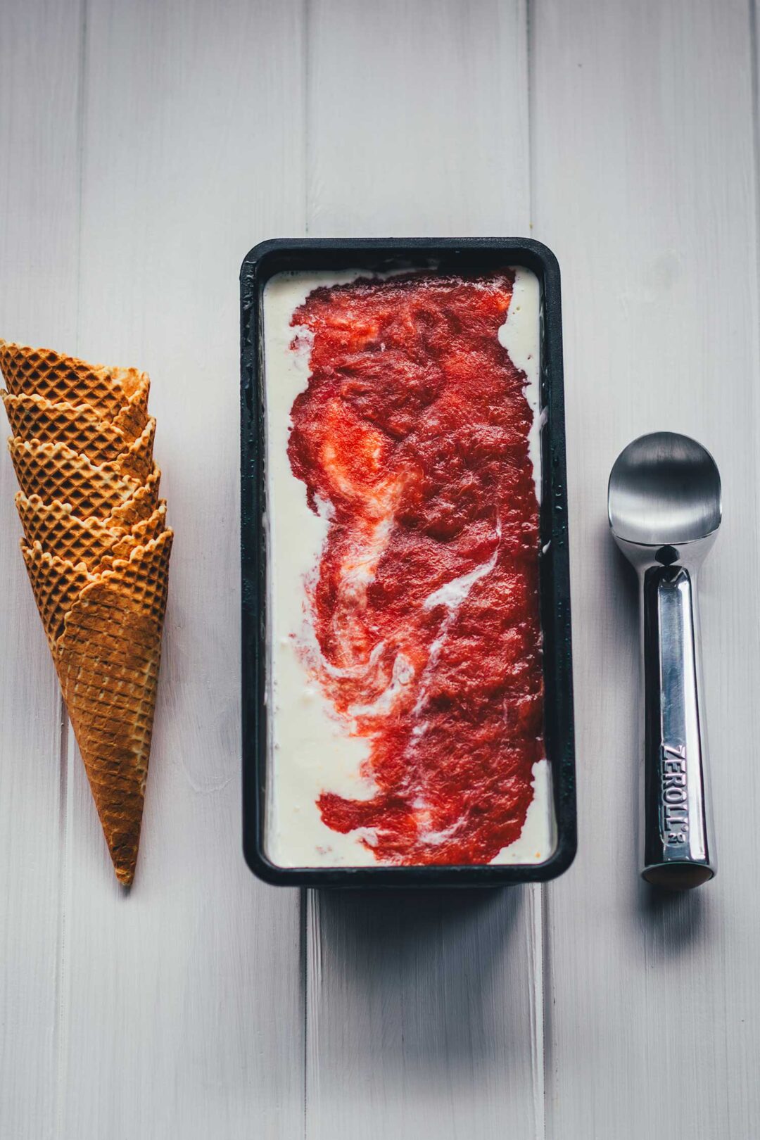 Selbstgemachtes Rhabarber-Eis mit Streuseln in einer schwarzen Eisform, daneben gestapelte Waffelhörnchen und ein Eisportionierer.
