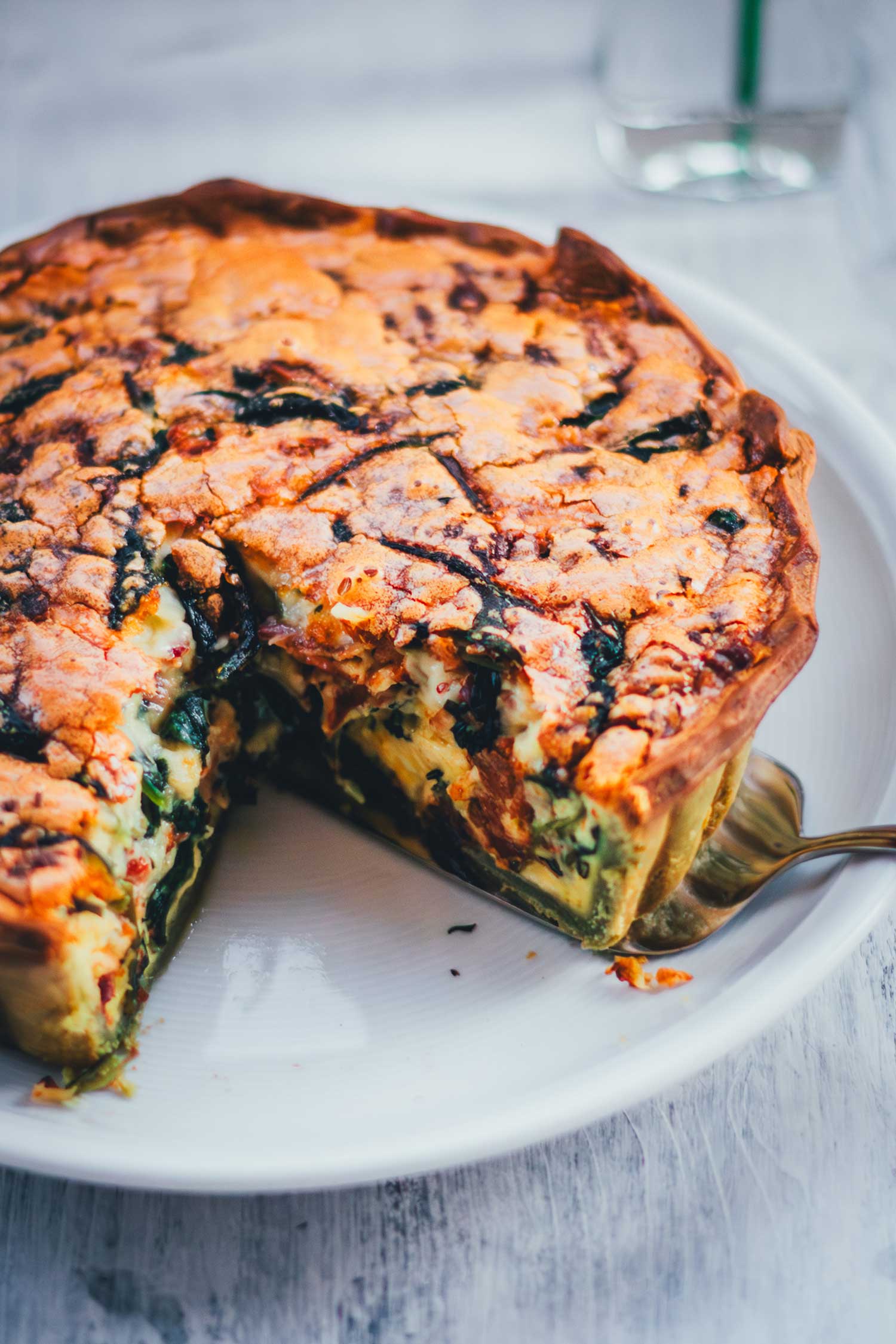 Seitliche Ansicht und Nahaufnahme einer frisch gebackenen Spinat-Bacon-Quiche mit Ricotta auf einem weißen Teller. Ein Stück wurde bereits herausgeschnitten, so dass man den Anschnitt mit der Füllung sieht: cremig und saftig!