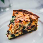 Nahaufnahme von einem einzelnen Stück Spinat-Bacon-Quiche auf einem weißen Teller. Man erkennt den grünen Spinat-Ricotta-Boden und die saftige Füllung der Quiche.