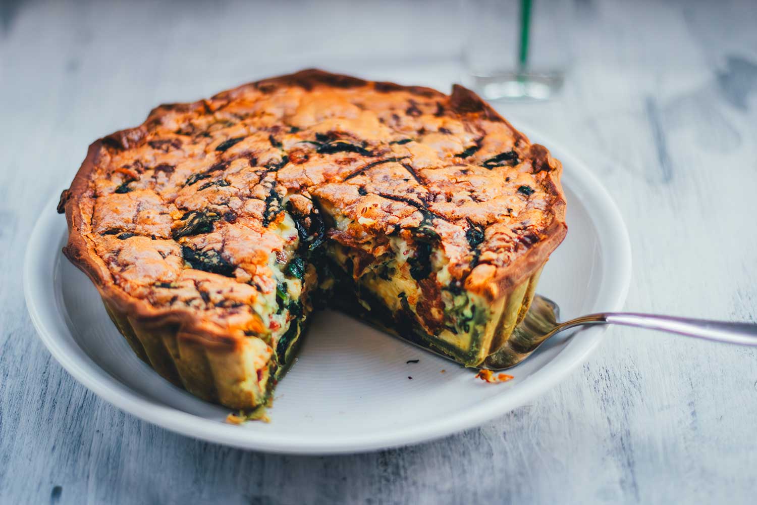 Spinat-Bacon-Quiche auf einem Teller serviert. Ein Stück wurde bereits herausgeschnitten und man erkennt den grünen Ricotta-Spinat-Boden und die würzige Füllung mit Bacon und Spinat.