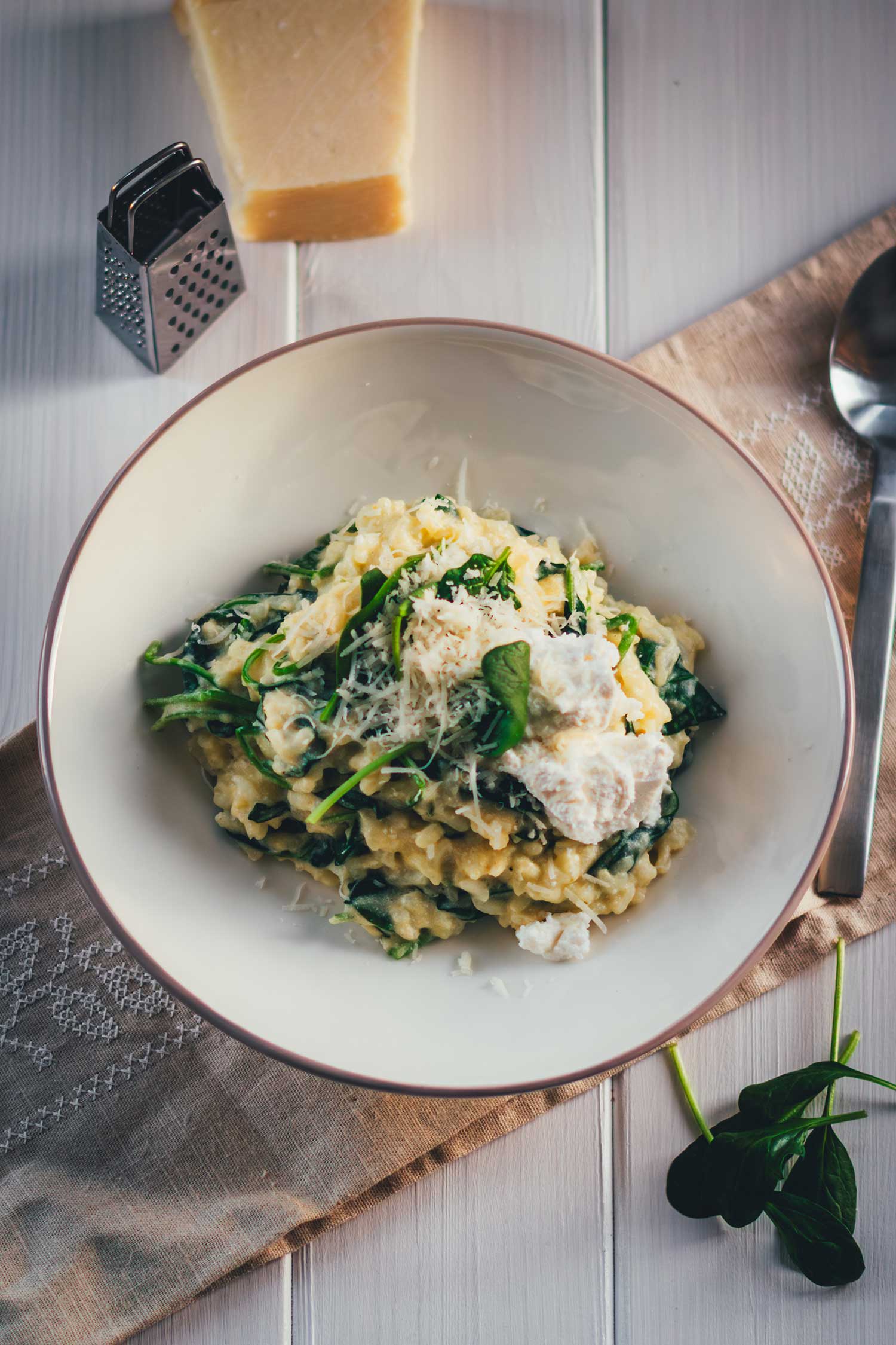 Topshot-Aufnahme von einem Teller mit Ofenrisotto. Man sieht Ricotta und frisch geriebenen Parmesan. Daneben liegt das parmesanstück mit Reibe, man sieht einen Esslöffel und etwas Babyspinat.