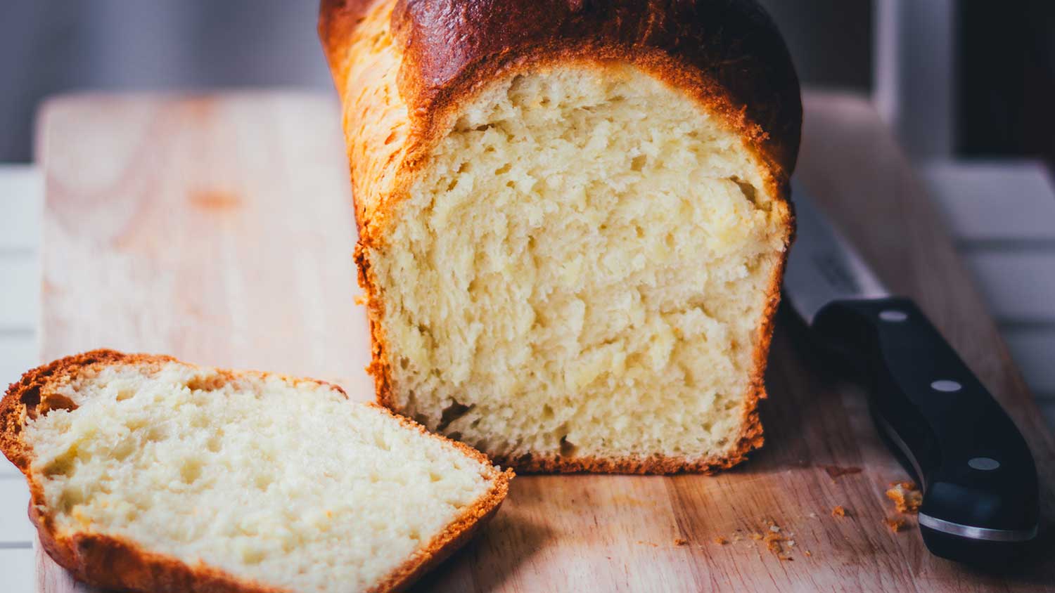 Nahaufnahme von einem frisch gebackenen und angeschnittenen Brioche-Brot auf einem Holzbrett. Man sieht die wattigweiche Krume und goldene Kruste. Daneben liegt ein Brotmesser.
