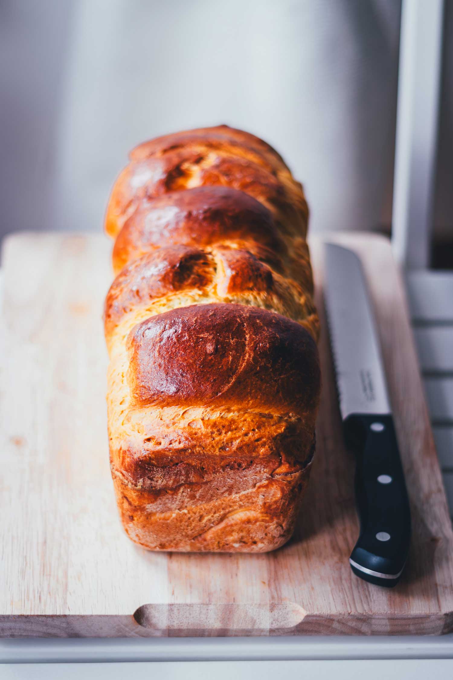 Frisch gebackenes Briochebrot aus einer Kastenform liegt goldbraun auf einem Holzbrett. Daneben liegt ein Brotmesser, bereit zum Anschneiden.