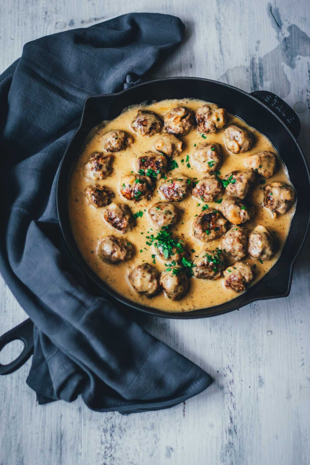 Köttbullar, schwedische Hackbällchen wie von IKEA werden in leckerer cremiger Sauce in einer gusseisernen Pfanne serviert. Die saftigen Hackbällchen sind mit Petersilie bestreut. Neben der Pfanne liegt ein Küchentuch. | moeyskitchen.com