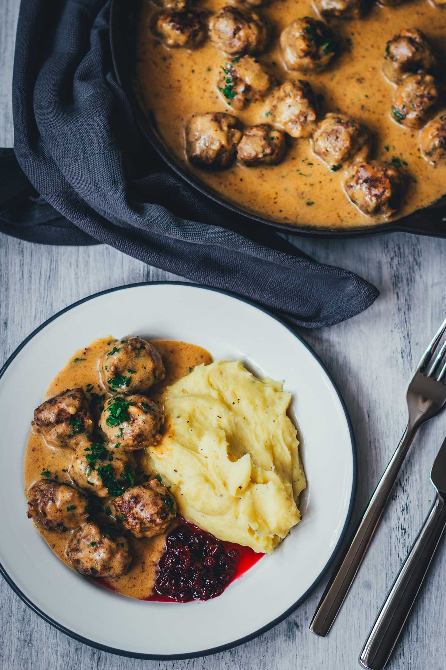 Topshotaufnahme eines fertig angerichteten Tellers mit schwedischen Hackbällchen, serviert mit Kartoffelbrei und Preiselbeeren. Im Hintergrund steht eine gusseiserne Pfanne, gefüllt mit Kötbullar in Sauce. | moeyskitchen.com