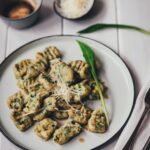 Seitliche, leicht schräge Draufsicht auf einen Teller mit frisch gekochten Bärlauch-Ricotta-Gnocchi. Serviert mit brauner Butter und geriebenem Käse im Hintergrund.