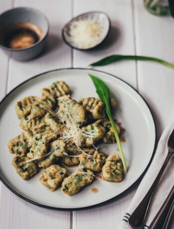 Seitliche, leicht schräge Draufsicht auf einen Teller mit frisch gekochten Bärlauch-Ricotta-Gnocchi. Serviert mit brauner Butter und geriebenem Käse im Hintergrund.