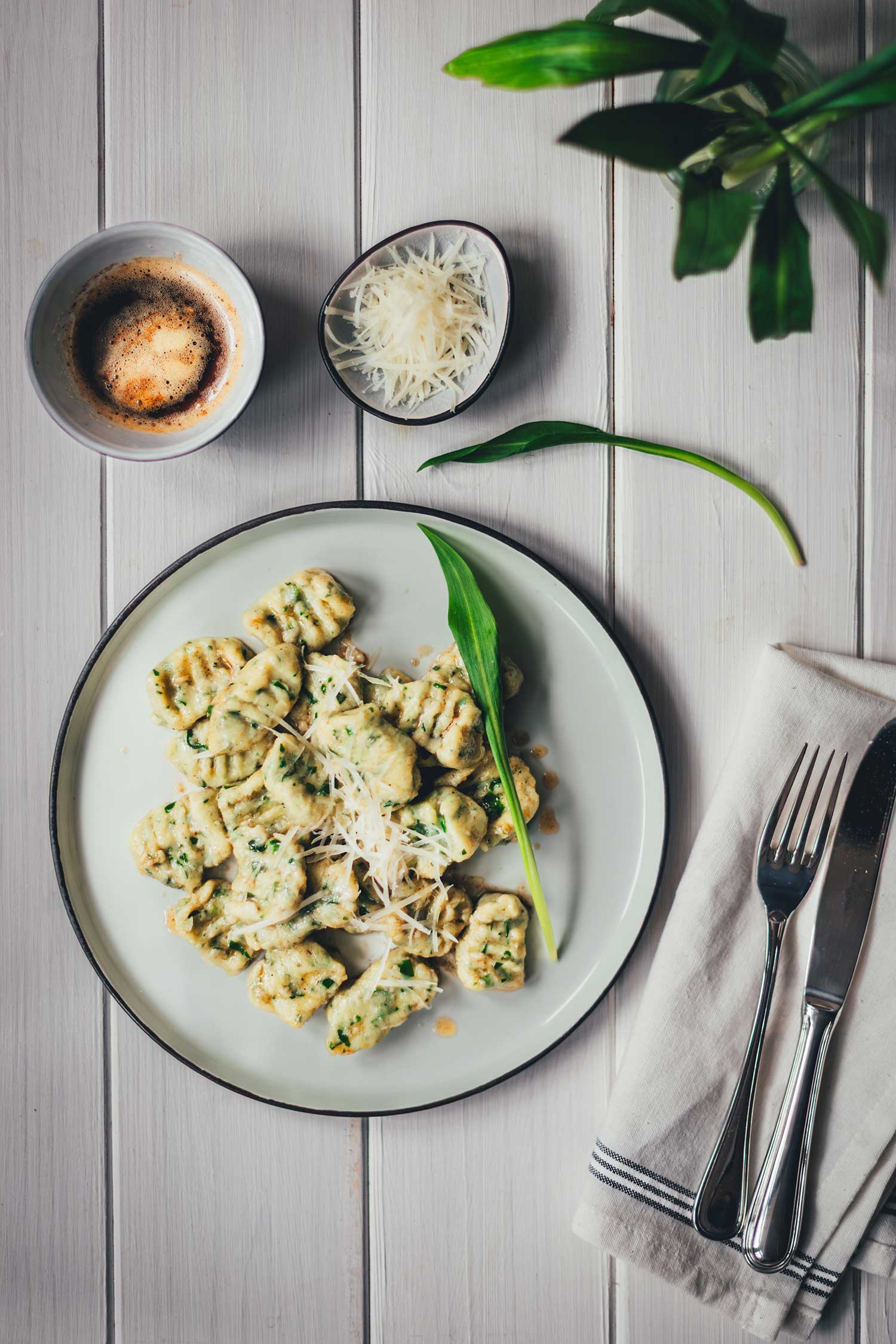 Draufsicht von oben auf einen Teller mit Bärlauch-Ricotta-Gnocchi, daneben liegen eine Serviette mit Besteckt, ein Bund Bärlauch sowie Schälchen mit brauner Butter und geriebenem Käse.