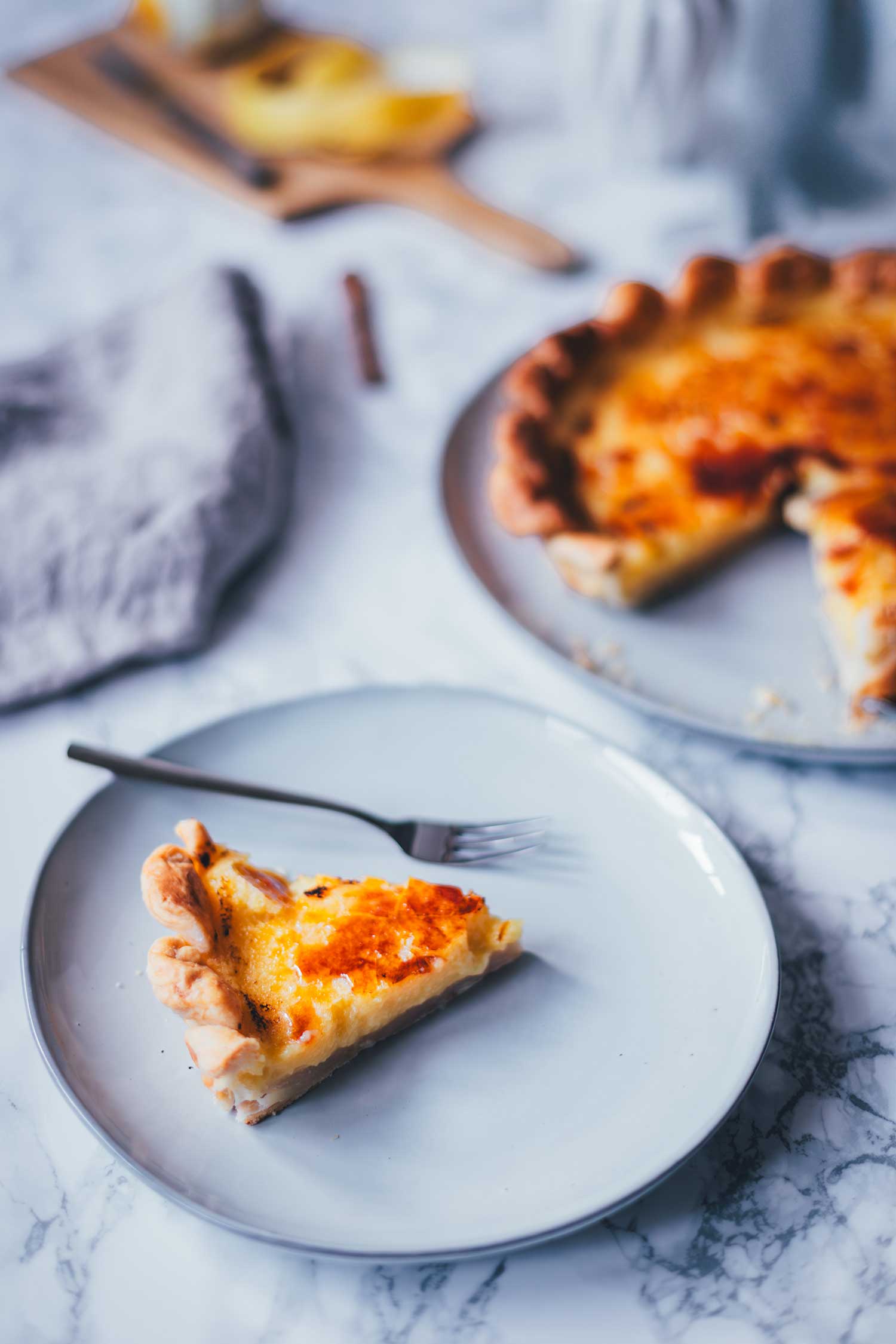Seitliche Ansicht einer frisch angeschnittenen pasteis de Nata Tarte. Ein Stück liegt auf einem grauen Teller, im Hintergrund sieht man die Zitronenschale.