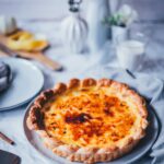 Seitliche Ansicht von einer großen Pasteis de Nata tarte auf einem runden Teller. Man erkennt die goldgelbe Füllung, den karamellisierten Zucker und den knusprigen Blätterteig. Im Hintergrund sieht man gelbe Blumen in weißen Vasen und einen marmorfarbenen Untergrund.