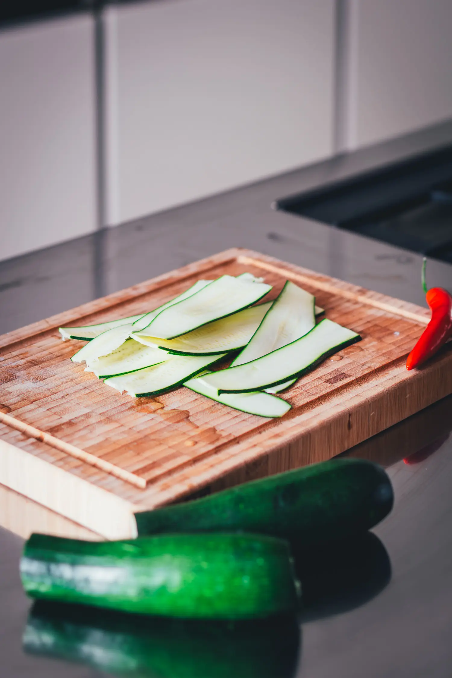 Dünn gehobelte Zucchinistreifen auf einem Holzbrett mit roter Chilischote, vorbereitet für den gegrillten Zucchini-Salat