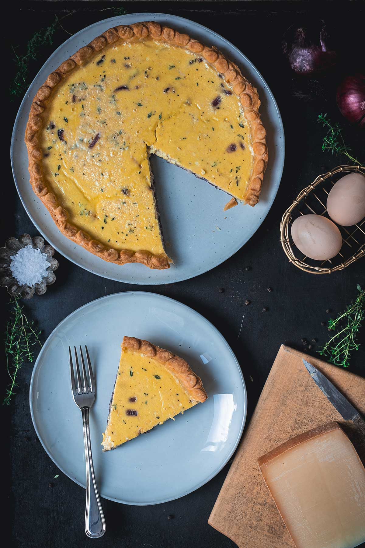 Zwiebelquiche von oben, angeschnitten mit einem Stück auf Teller serviert, dekoriert mit Thymian und ganzen Zwiebeln, Eiern und Bergkäse | moeyskitchen.com