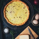 Zwiebelquiche von oben fotografiert, ganze Quiche mit karamellisierten roten Zwiebeln, daneben Thymian, Eier, Bergkäse, Salz und Zwiebeln als Deko | moeyskitchen.com