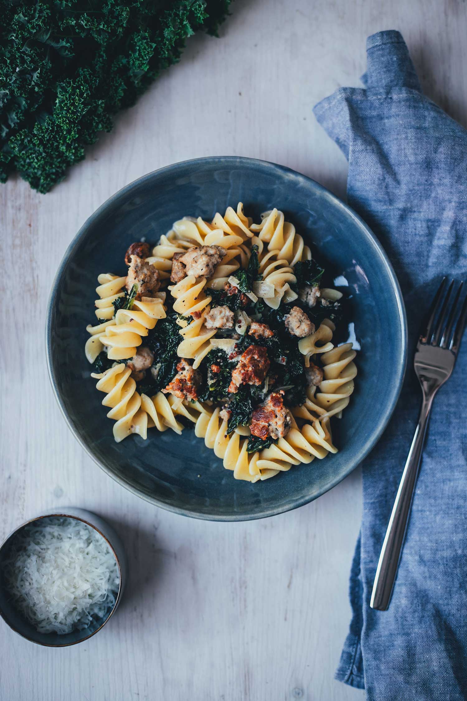 Topshotaufnahme eines dunkelblauen Tellers, gefüllt mit Pasta mit Grünkohl und Bratwurstbrät und Senfsauce. Daneben liegt eine kleine Schüssel mit geriebenem Parmesan, einige Blätter Grünkohl und eine hellblaue Serviette mit einem Esslöffel