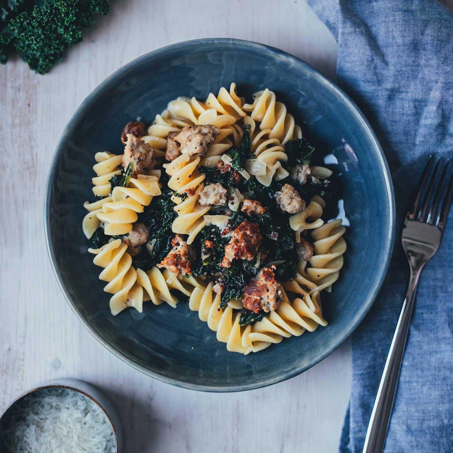 Topshotaufnahme eines dunkelblauen Tellers, gefüllt mit Pasta mit Grünkohl und Bratwurstbrät und Senfsauce. Daneben liegt eine kleine Schüssel mit geriebenem Parmesan, einige Blätter Grünkohl und eine hellblaue Serviette mit einem Esslöffel