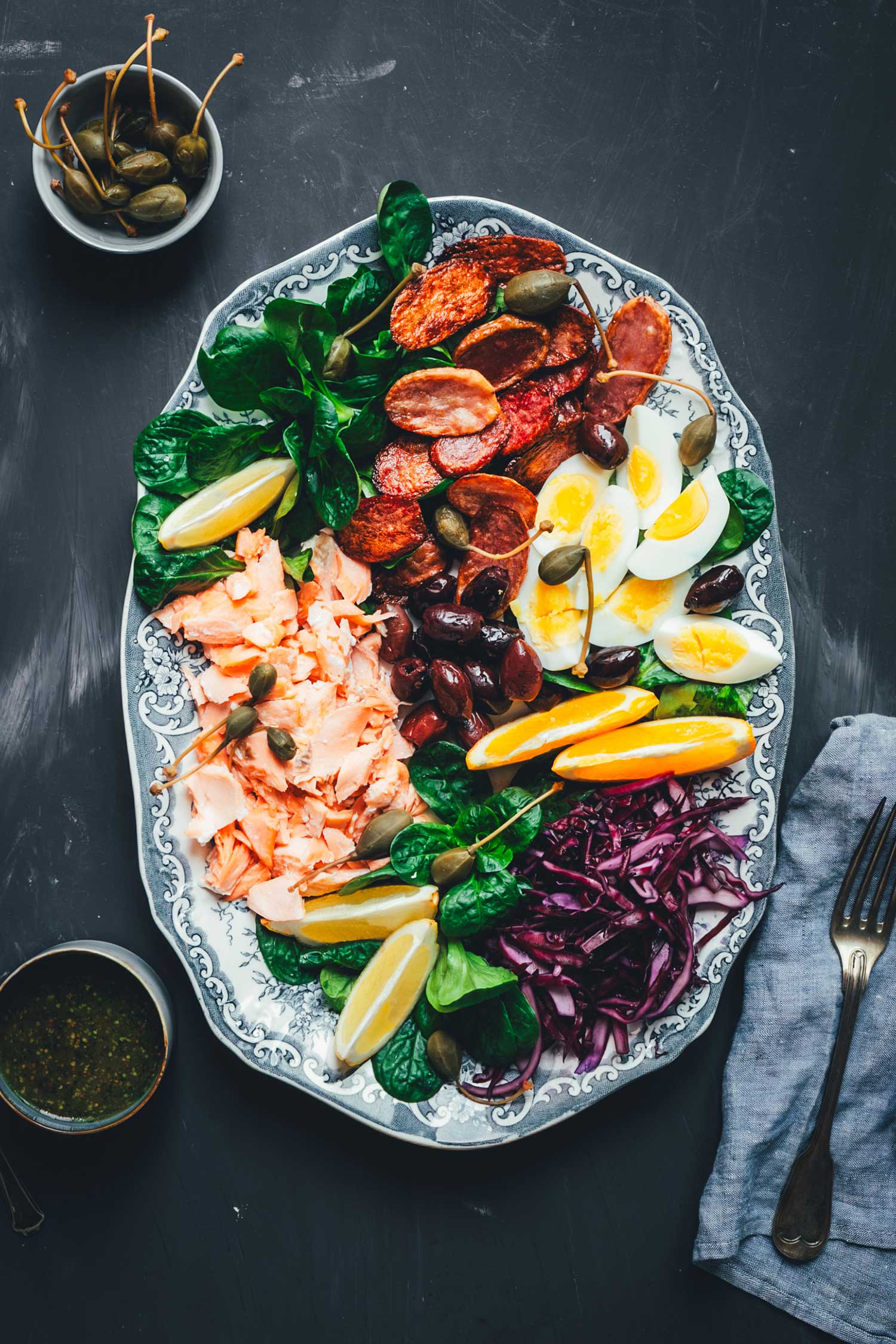 Topshot Winterlicher Nizza Salat mit Pulled Lachs auf Servierplatte – mit Rotkohl, Feldsalat, knusprigen Kartoffeln, Eiern, Orangen und Kapernäpfeln. Daneben das Dressing in einer kleinen Schale.