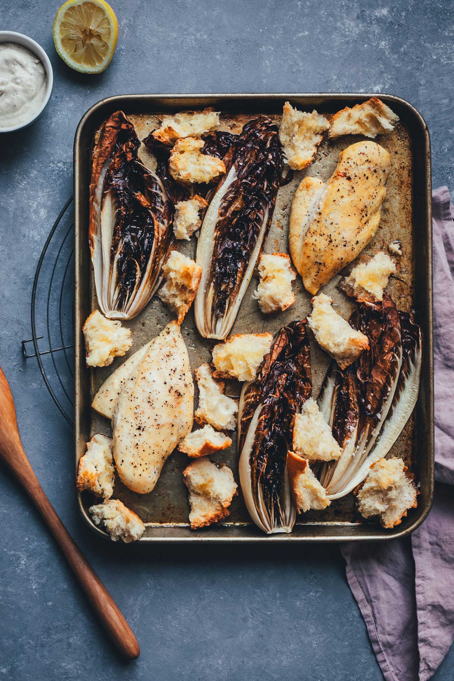 Draufsicht auf das fertige Blechgericht direkt nach dem Backen: Saftige Hähnchenbrustfilets, geröstete Radicchio-Viertel und knusprige Brotstücke auf einem dunklen Backblech.