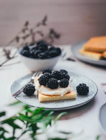 Saftiges Stück Hot Milk Sponge Cake, üppig mit Mascarpone-Creme und frischen Brombeeren belegt, auf einem gesprenkelten Teller.