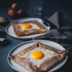 Seitliche Ansicht von zwei Tellern mit frisch gebackenen Galettes Complètes – französischen Buchweizenpfannkuchen aus der Bretagne, die mit Käse, Schinken und Spiegelei gefüllt sind.