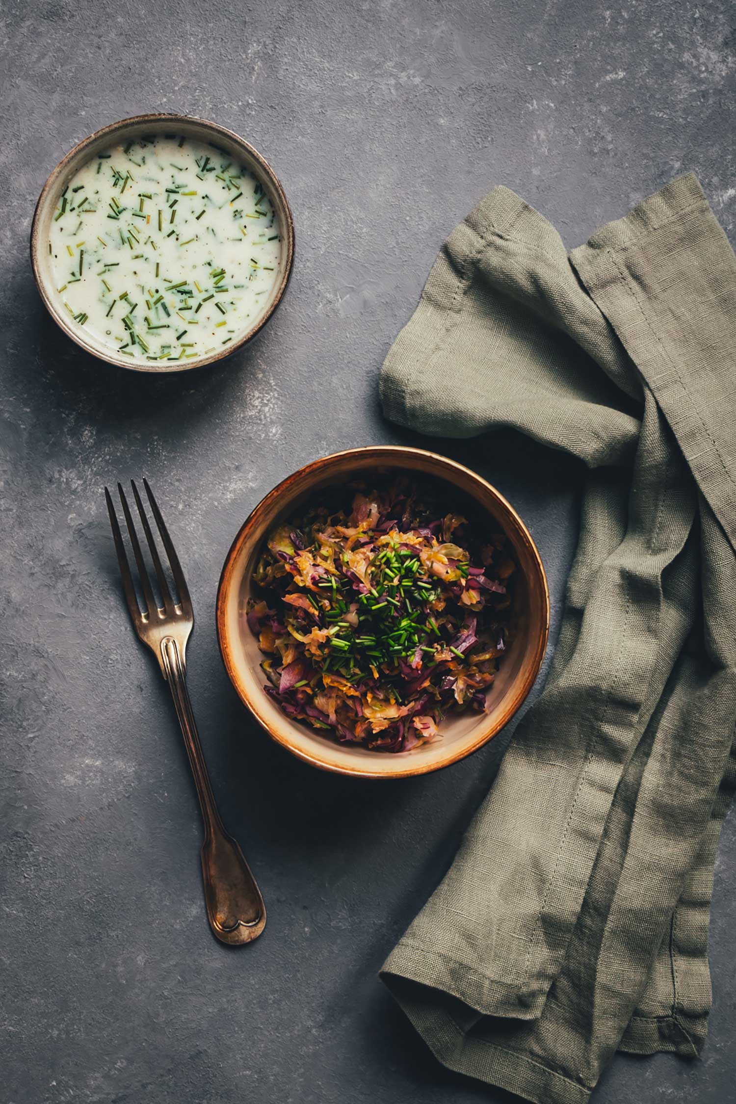 Top-down: Bunter Krautsalat in einer Schale mit Schnittlauch, daneben Gabel und Leinentuch – oben eine Schale Buttermilch-Dressing.