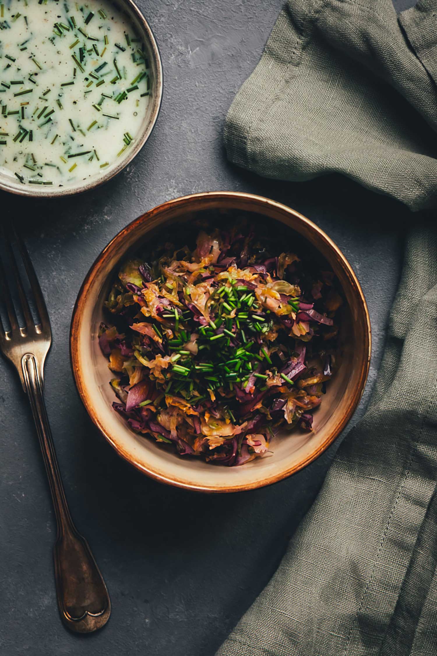 Nahaufnahme von Krautsalat mit Schnittlauch in einer Keramikschale, daneben Gabel und Leinentuch – oben eine Schale Buttermilch-Zitronen-Dressing.