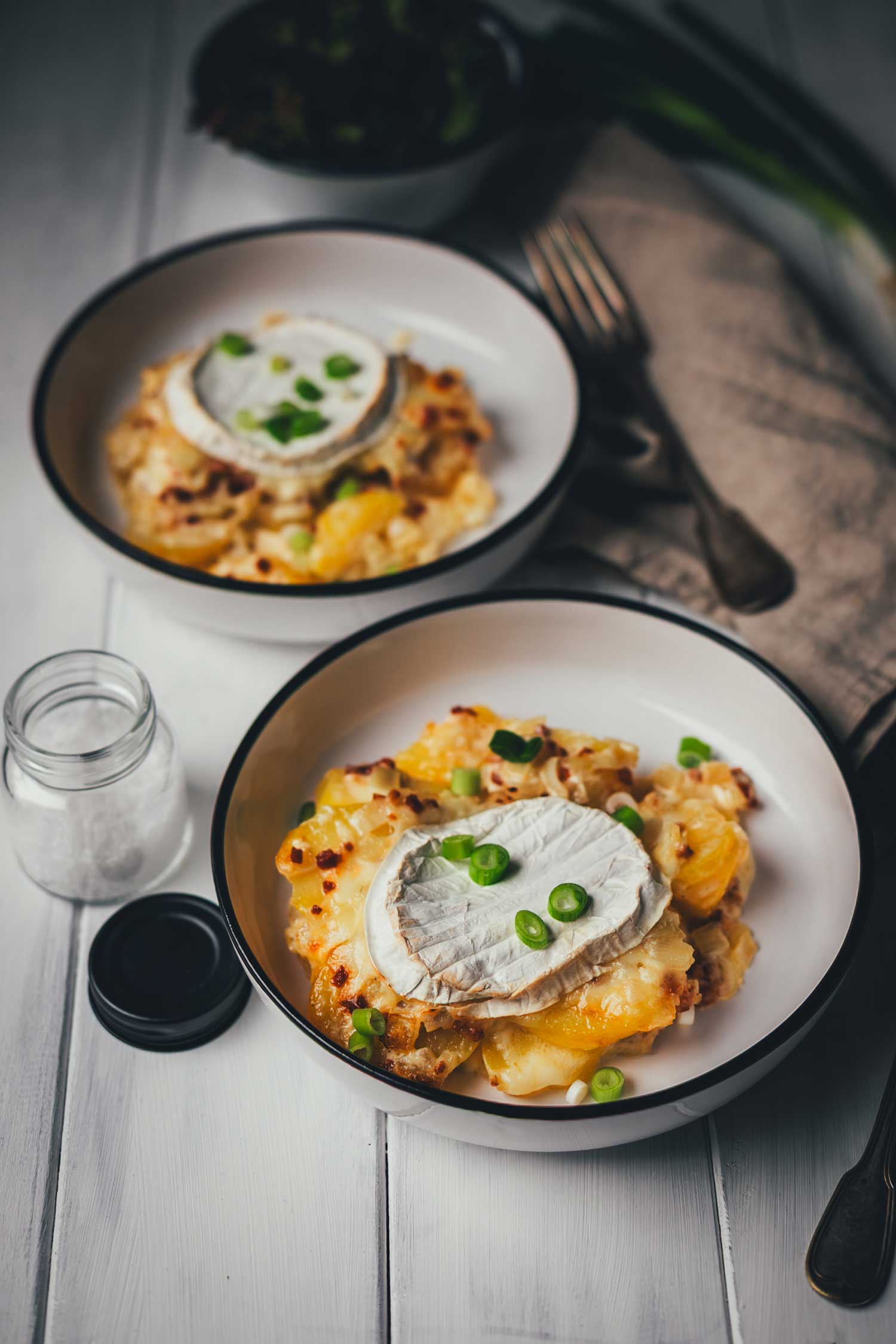 * Zwei servierte Portionen Tartiflette mit Camembert in weißen Schalen auf einem hellen Holztisch, garniert mit frischen Frühlingszwiebeln, daneben eine Leinenserviette und Besteck.