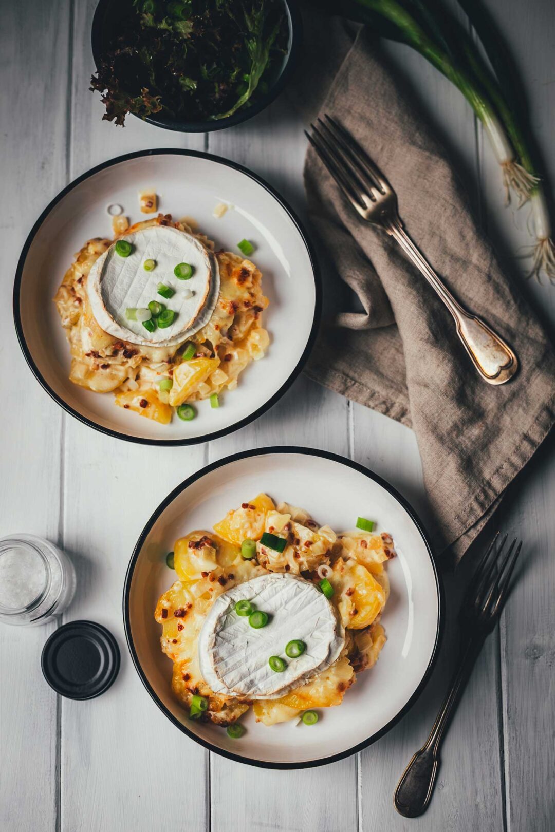 Draufsicht auf einen gedeckten Tisch mit zwei Portionen Tartiflette mit Camembert in weißen Schüsseln, einer Schale mit grünem Salat und einer Leinen-Serviette auf hellem Holzgrund.