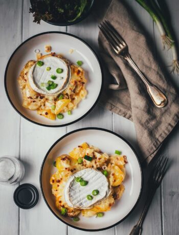 Draufsicht auf einen gedeckten Tisch mit zwei Portionen Tartiflette mit Camembert in weißen Schüsseln, einer Schale mit grünem Salat und einer Leinen-Serviette auf hellem Holzgrund.