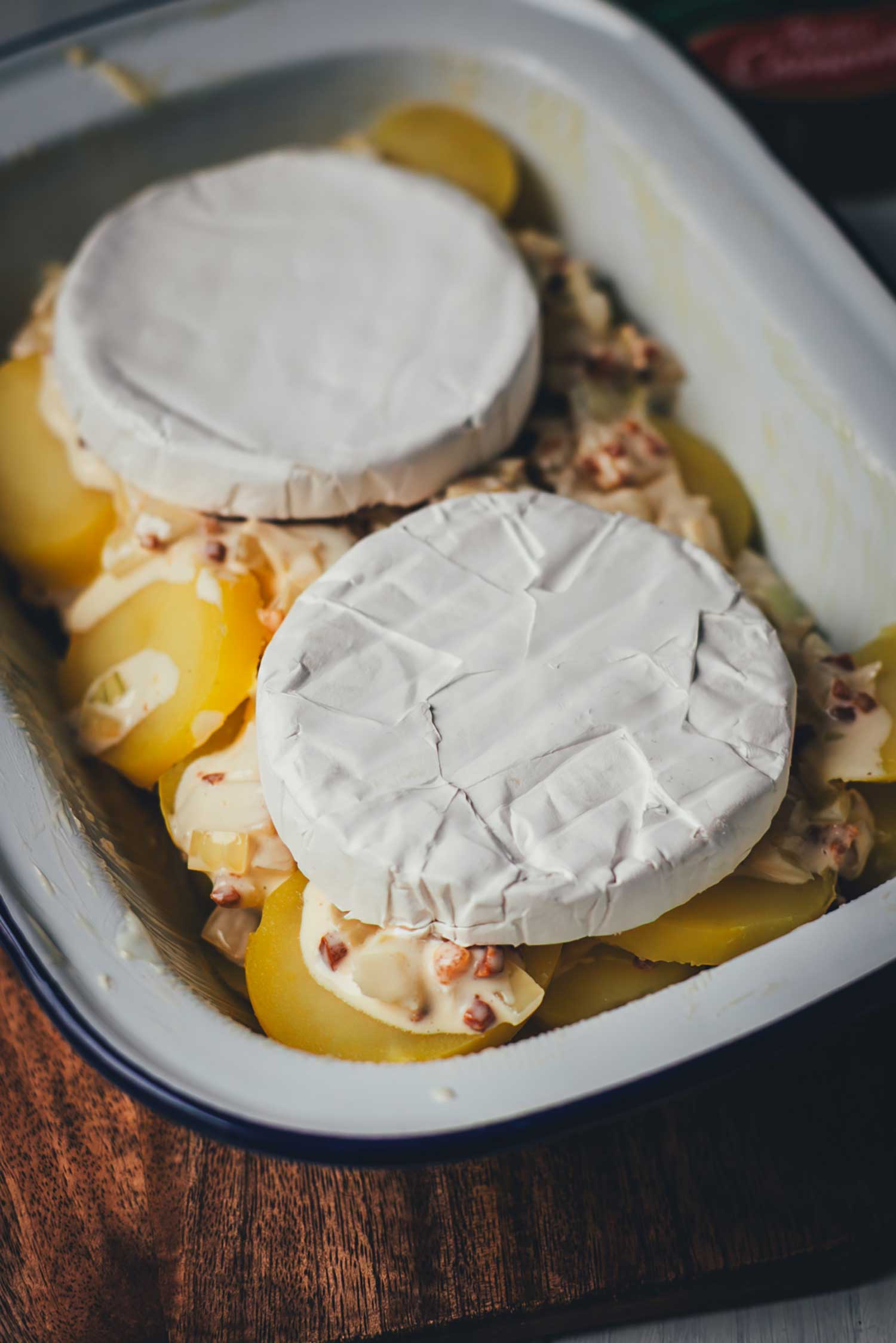 Vorbereitung einer Tartiflette mit Camembert: halbierte Camembert-Laibe liegen mit der Rinde nach oben auf einer Schicht aus Kartoffelscheiben, Speck und Zwiebeln in einer hellen Auflaufform.