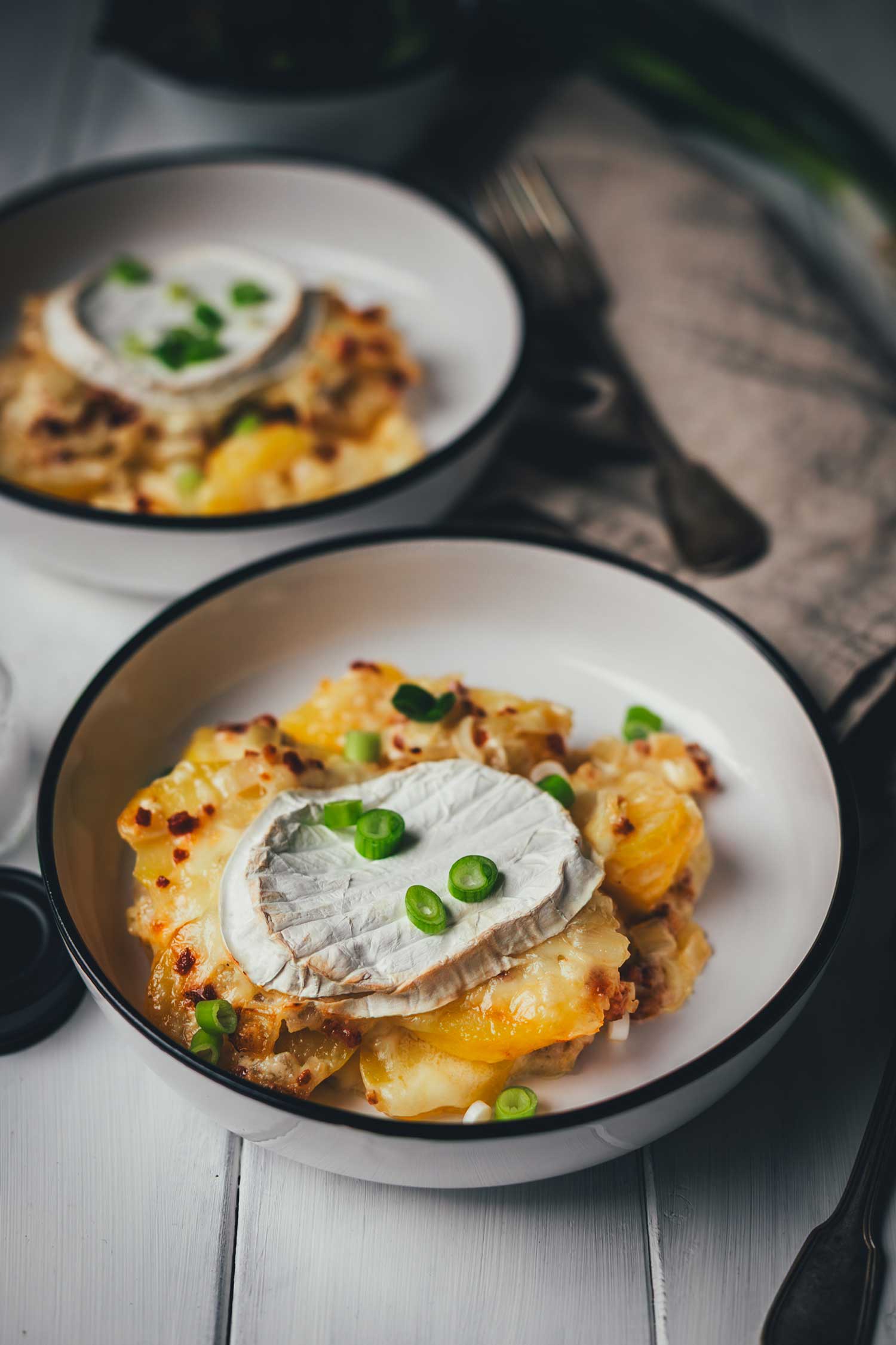 Eine Portion fertige Tartiflette mit Camembert auf einem weißen Teller angerichtet, garniert mit frischen Frühlingszwiebelringen. Im Hintergrund ist eine zweite Portion erkennbar.