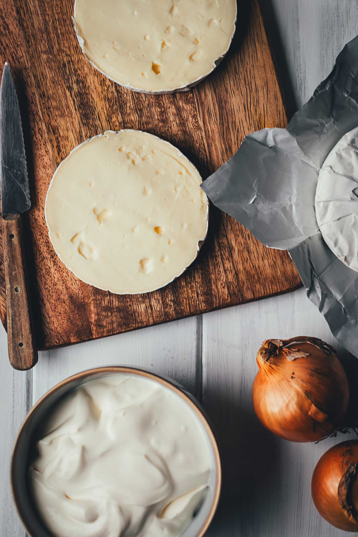 Zutaten für eine Tartiflette mit Camembert auf einem hellen Holztisch: Ein waagerecht halbierter Camembert auf einem Holzbrett, eine Schale mit Crème fraîche und frische Zwiebeln.