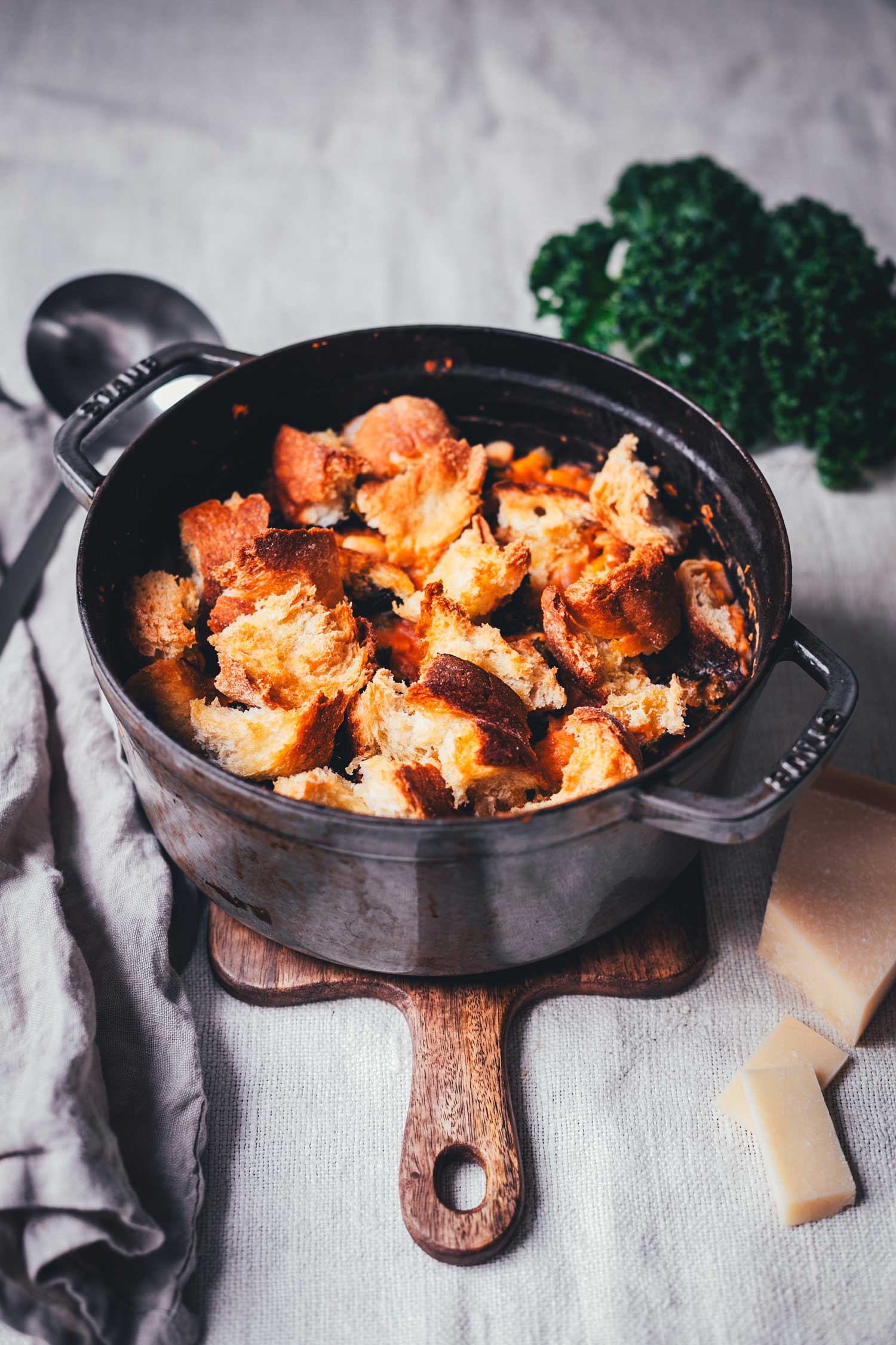Seitliche Ansicht von einem gusseisernen Topf, gefüllt mit Eintopf und getoppt mit Brot. Daneben sieht man eine Suppenkelle, ein Küchentuch, etwas Grünkohl und Parmesan | moeyskitchen.com
