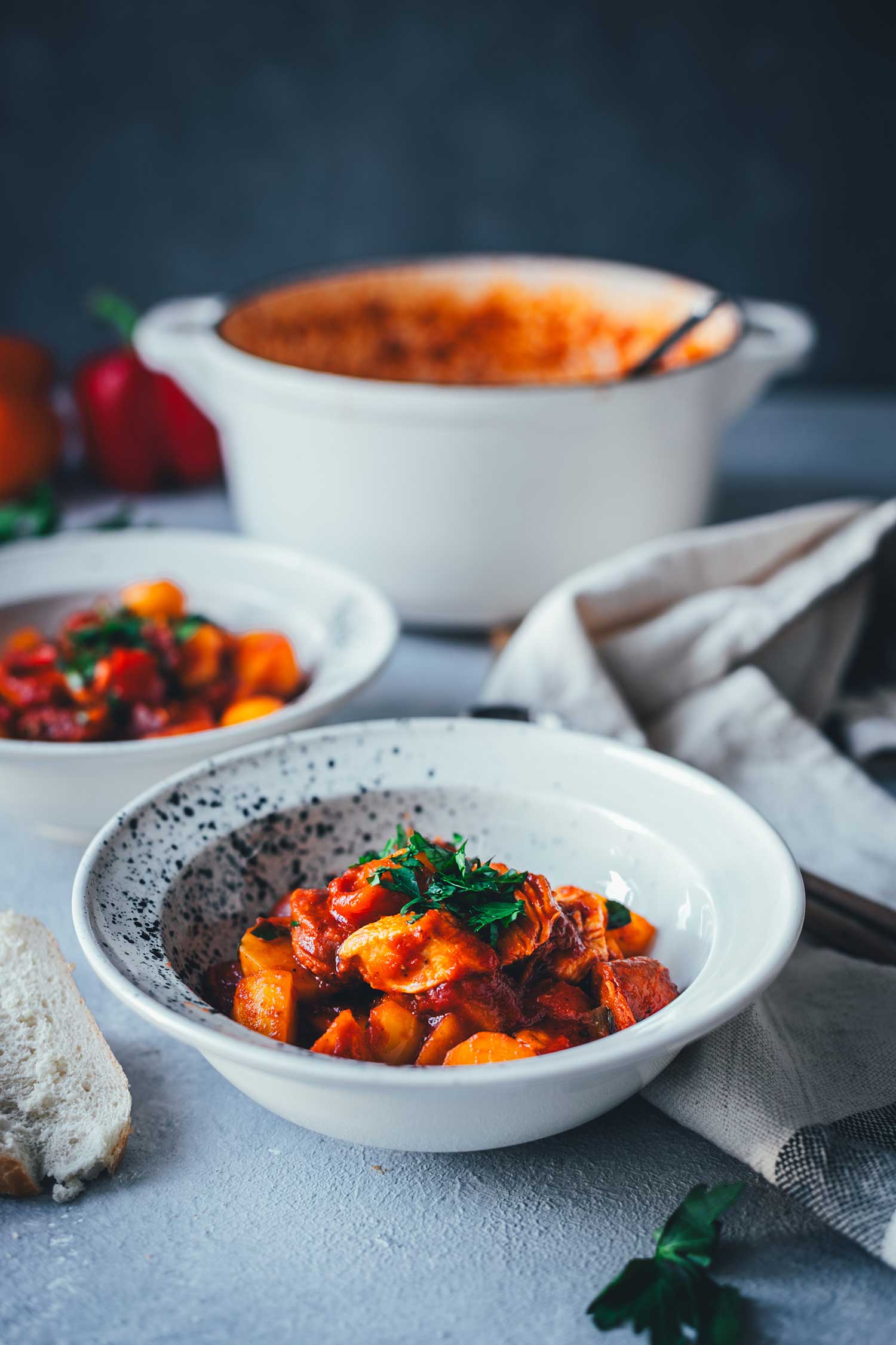 Seitliche Nahaufnahme von einer Schüssel mit einem deftigen Eintopf aus Kartoffeln, Paprika, Chorizo und Hähnchenbrust. Im Hintergrund sieht man den Schmortopf und einen weiteren Teller. | moeyskitchen.com