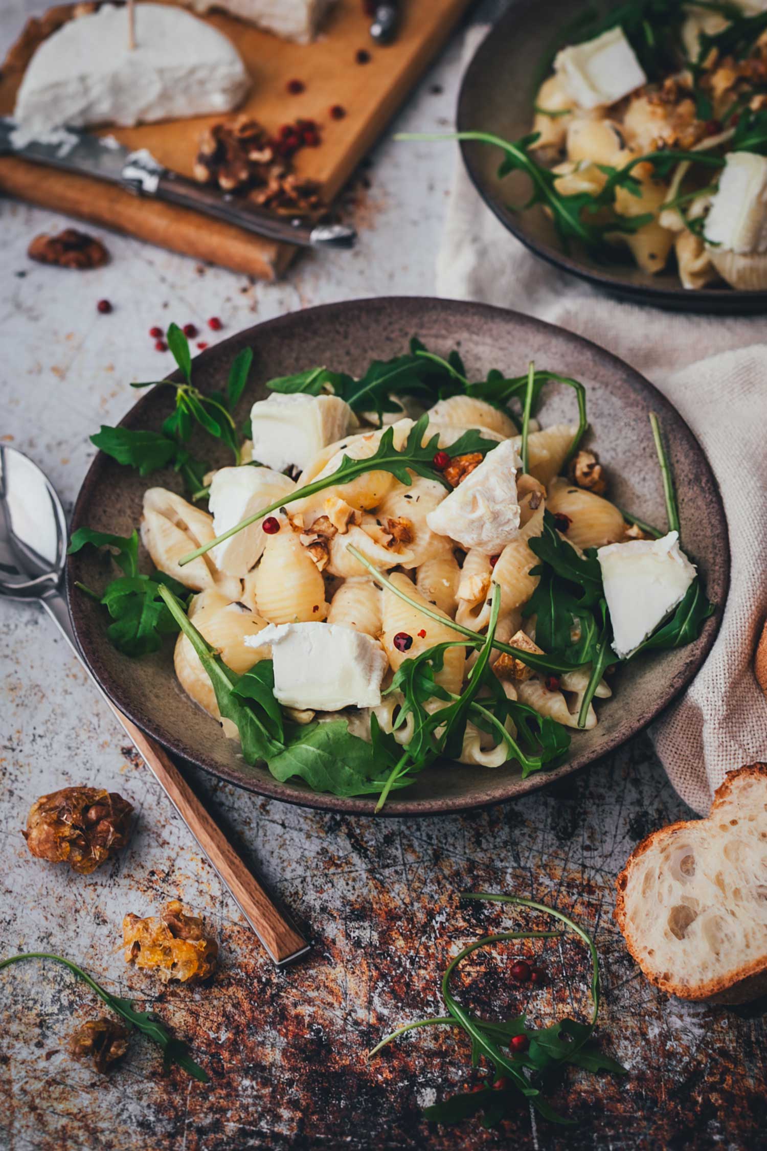 Leicht erhöhte Draufsicht auf einen gerade servierten tiefen Teller mit Pasta. Muschelnudeln werden mit Ziegenkäse-Sauce, Ziegenkäse, Walnüssen und Rucola zu etwas Baguette serviert.